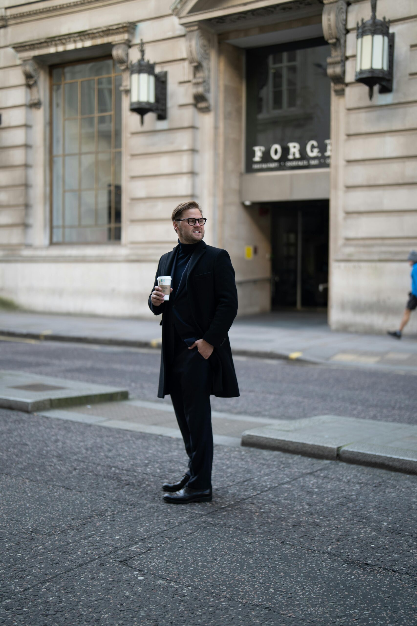 A man in a black suit stands with a coffee cup on a city street, blending modern fashion with urban life.